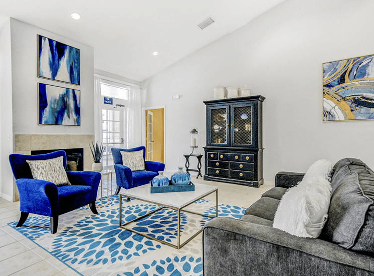 Clubhouse furniture with gray couch and white pillows, blue fabric chairs with pillows, a cabinet in the background with decor and abstract art on walls